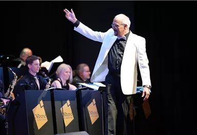 Ein Dirigent in einem weißen Smoking leitet ein Orchester. Im Hintergrund sind Musiker mit ihren Instrumenten zu sehen.