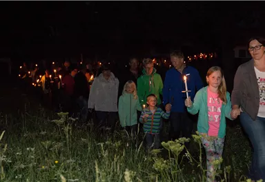 Eine Gruppe von Menschen geht bei Nacht, alle mit Kerzen in der Hand. Im Hintergrund sind Bäume und Wiese zu sehen.