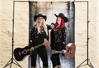 Zwei Musikerinnen stehen vor einem Hintergrund und halten Gitarren. Sie tragen schwarze Outfits und Hüte.