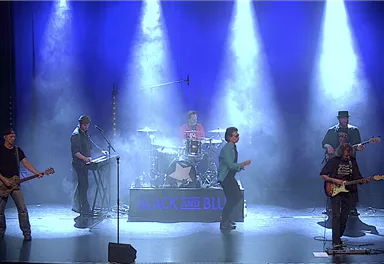 Eine Band spielt live auf der Bühne mit farbigem Licht und Nebel. Die Musiker sind in lebhaften Posen und nutzen verschiedene Instrumente.