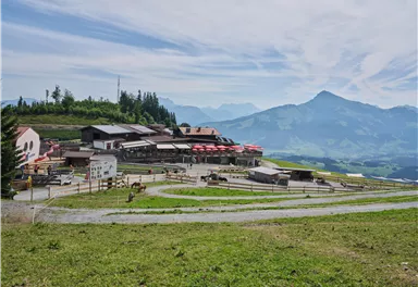Eine malerische Berglandschaft mit einem rustikalen Gebäude und roten Sonnenschirmen. Im Hintergrund sind majestätische Berge und ein klarer Himmel zu sehen.