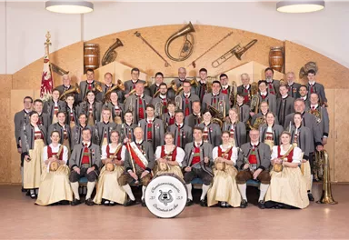 Eine große Gruppe von Musikern in traditionellen Trachten steht zusammen. Im Hintergrund sind Musikinstrumente und eine Fahne sichtbar.