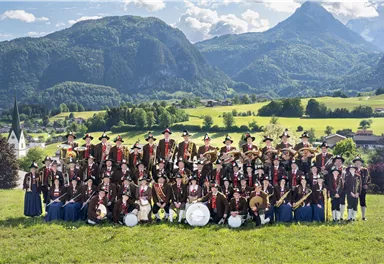 Eine große Gruppe von Menschen steht in traditioneller Tracht auf einer Wiese, umgeben von Bergen. Im Hintergrund sind grüne Felder und ein blauer Himmel zu sehen.