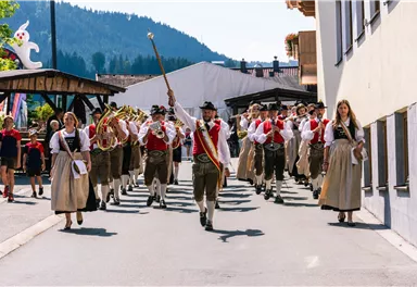 A traditional parade with people in traditional costumes walking down a street. In the background, mountains and festive stalls can be seen.
