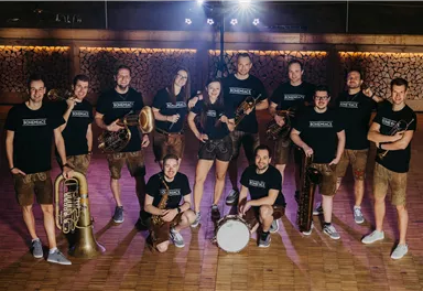 Eine Gruppe von Musikern in traditionellen Trachten steht auf einer Holzfläche. Sie halten verschiedene Musikinstrumente und posieren gemeinsam für das Foto.
