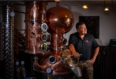 A man stands next to a large copper distillery in a distillery. The machine has various pressure gauges and a shiny surface.