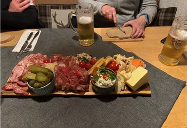 Ein reichhaltiges Brett mit verschiedenen Wurst- und Käsesorten, eingelegten Gurken und frischen Tomaten. Dazu zwei Biere auf einem Tisch.