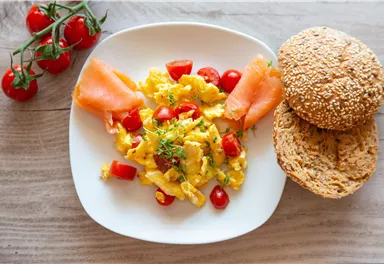 A plate with scrambled eggs, cherry tomatoes, and smoked salmon. Next to it are two sesame rolls.