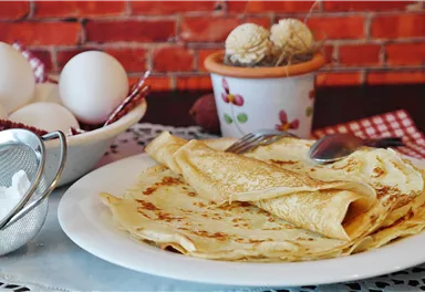 A plate of fresh crêpes is on a table. Next to it are eggs and a container of powdered sugar.