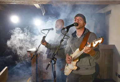 Zwei Musiker treten auf einer Bühne auf und spielen akustische Gitarre. Nebel und Scheinwerferlicht schaffen eine stimmungsvolle Atmosphäre.