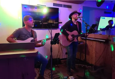 Ein Musiker spielt Gitarre und singt live, während ein anderer Musiker am Bass begleitet. Die Umgebung ist mit buntem Licht beleuchtet.