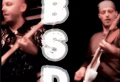 Two musicians are playing guitar on a stage. The background is dark and the letters "B S D" are prominently placed.