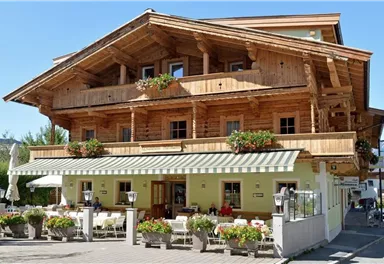 A traditional wooden house with a balcony and flower boxes. The entrance area is inviting with tables and chairs outside.