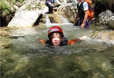 A child is swimming in clear water wearing a red helmet. In the background, other people and rocks can be seen.