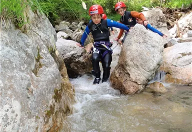 Two children in wetsuits are climbing over rocks in a stream. They are wearing helmets and look focused as they explore the water.