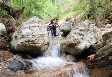 A group of people is crossing a small river, surrounded by large stones and dense green vegetation. The scene conveys a sense of adventure in nature.