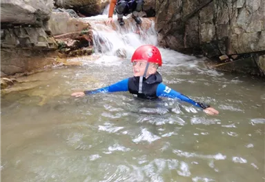 A boy swims in clear, flowing water and wears a red helmet. In the background, rocks and a small waterfall can be seen.