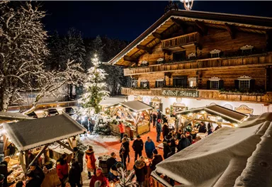 A festive winter market scene with people visiting stalls. In the background stands a rustic wooden house, surrounded by snow-covered trees and a lit Christmas tree.