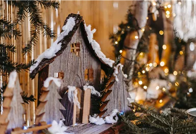 A decorative wooden house with snow and small fir trees. In the background, lights and festive decorations can be seen.