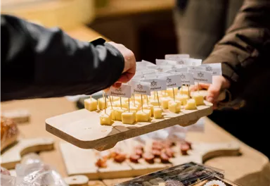 A hand is holding a wooden platter with small cheese samples, each labeled with little flags. In the background, more delicacies can be seen on a table.