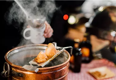 A steaming pot of fried foods is being prepared. In the background, blurred silhouettes of people can be seen.