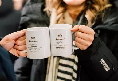 Two cups clink together, held by people in warm clothing. In the background, a blurred, dark environment is visible.
