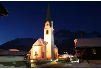 A charming church with a tall, green tower, surrounded by snow. The scene is characterized by a clear night sky and gentle lighting of the buildings.