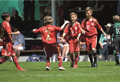 Eine Gruppe von jungen Fußballspielern feiert ein Tor auf dem Spielfeld. Sie tragen rot-weiße Trikots und wirken glücklich und aufgeregt.