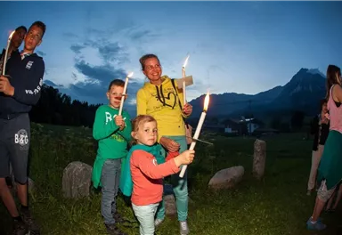 Eine Gruppe von Kindern und Erwachsenen hält Kerzen in einer ländlichen Umgebung bei Dämmerung. Im Hintergrund sind Berge und eine Wiese sichtbar.