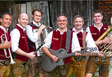 Sechs Musiker in traditionellen Trachten stehen vor einer Holzwand. Sie halten verschiedene Instrumente und lächeln in die Kamera.
