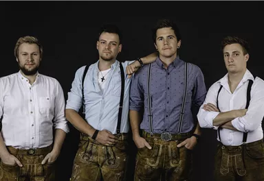 Four men in traditional Bavarian attire stand against a black background. They wear lederhosen and shirts, striking a confident pose.