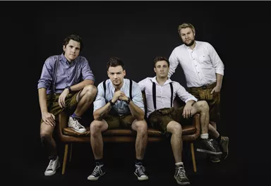 Four men in traditional Bavarian attire are sitting on a bench. They are wearing shirts, suspenders, and shoes, while the background is dark.