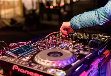 Ein DJ-Pult mit einem DJ, der an den Reglern und Knöpfen arbeitet. Bunte Lichter und eine festliche Atmosphäre sind im Hintergrund sichtbar.