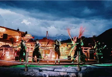 A group of performers presents a performance under dramatic lighting. In the background, buildings and a cloudy sky are visible.