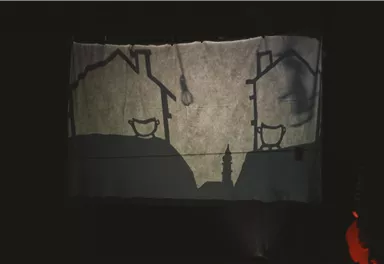 A shadow projection on a canvas shows two silhouetted houses. The scene appears mystical and dark, with some hinted objects in the foreground.