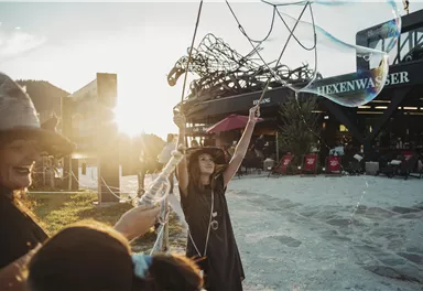 A woman with a hat catches large soap bubbles in the evening sun. In the background, a building with the inscription "Hexenwasser" can be seen.