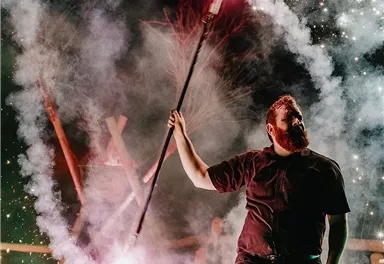 An artist strikes sparks with a torch, surrounded by smoke and fiery effects. The scene radiates tension and dynamism.