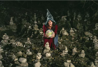 A mystical woman is wearing a magic hat and holding a glowing sphere. Around her, there are many stone turrets that create a mysterious atmosphere.