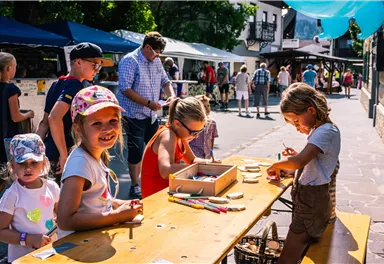 A cheerful market day with children crafting at the table. In the background, there are stalls and smiling visitors.