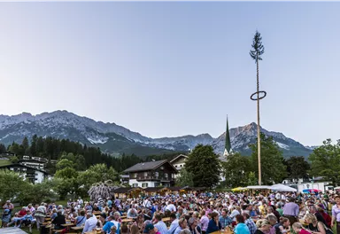 Eine große Menschenmenge versammelt sich in einem malerischen Bergdorf. Im Hintergrund sind die Berge und eine Kirche zu sehen, während ein Maibaum aufgestellt wird.