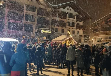 Eine große Menschenmenge steht in einem verschneiten Wintermarkt. Schnee fällt dicht über die festlich beleuchteten Gebäude.