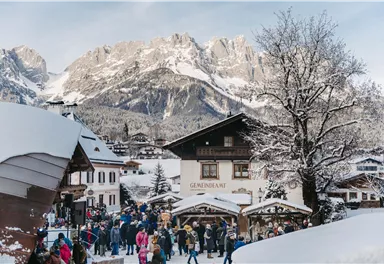 Ein verschneites Dorf mit vielen Menschen und traditionellen Gebäuden. Die beeindruckenden Berge im Hintergrund verleihen der Szene eine malerische Atmosphäre.