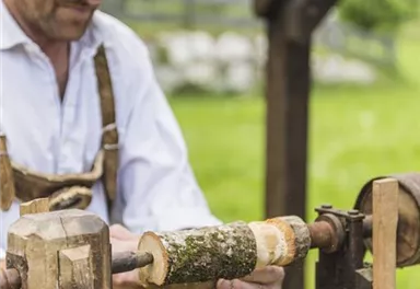 Ein Handwerker arbeitet an einer Drechselmaschine im Freien. Im Hintergrund sind grüne Wiesen und Bäume zu sehen.