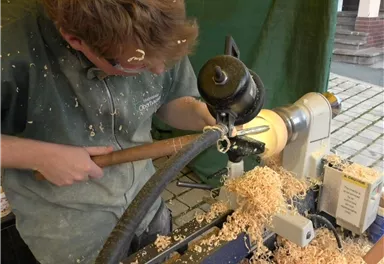 Ein Handwerker arbeitet an einer Drehmaschine und formt Holz. Späne fliegen herum und der Arbeitsplatz ist mit Holzspänen bedeckt.