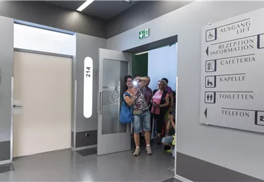 Eine Gruppe von Menschen steht am Eingang zu einem Flur in einem modernen Gebäude. Auf der Wand sind Wegweiser zu verschiedenen Einrichtungen wie Cafeteria und Toiletten.