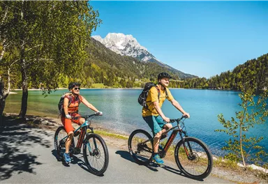 Two cyclists ride along a clear blue lake, surrounded by mountains and trees. The sun is shining and the sky is blue.