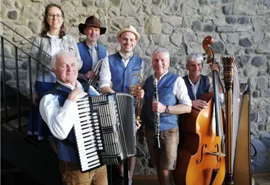 Sechs Musiker in traditionellen Kleidern stehen vor einer steinernen Wand. Sie halten verschiedene Instrumente wie Akkordeon, Klarinette und Harfe.
