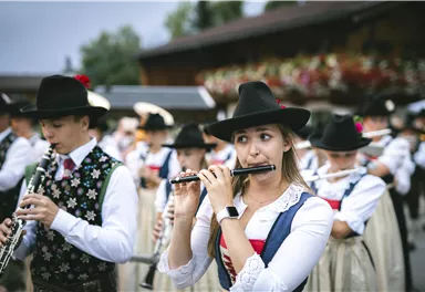 A chapel in traditional attire plays festive music. It marches down a picturesque street with green surroundings and mountains in the background.