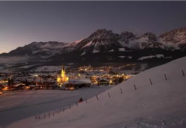 Eine malerische alpine Landschaft bei Nacht mit schneebedeckten Bergen. Im Vordergrund ist ein kleines Dorf mit beleuchteten Häusern und einer Kirche sichtbar.
