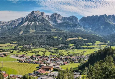 An impressive mountain landscape with green meadows and a clear sky. In the foreground, there are small houses and bustling fields.